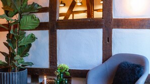Une pièce avec des poutres apparentes, un pouf rond, une chaise avec un coussin foncé, une plante en pot et une petite table avec des bougies et un vase.