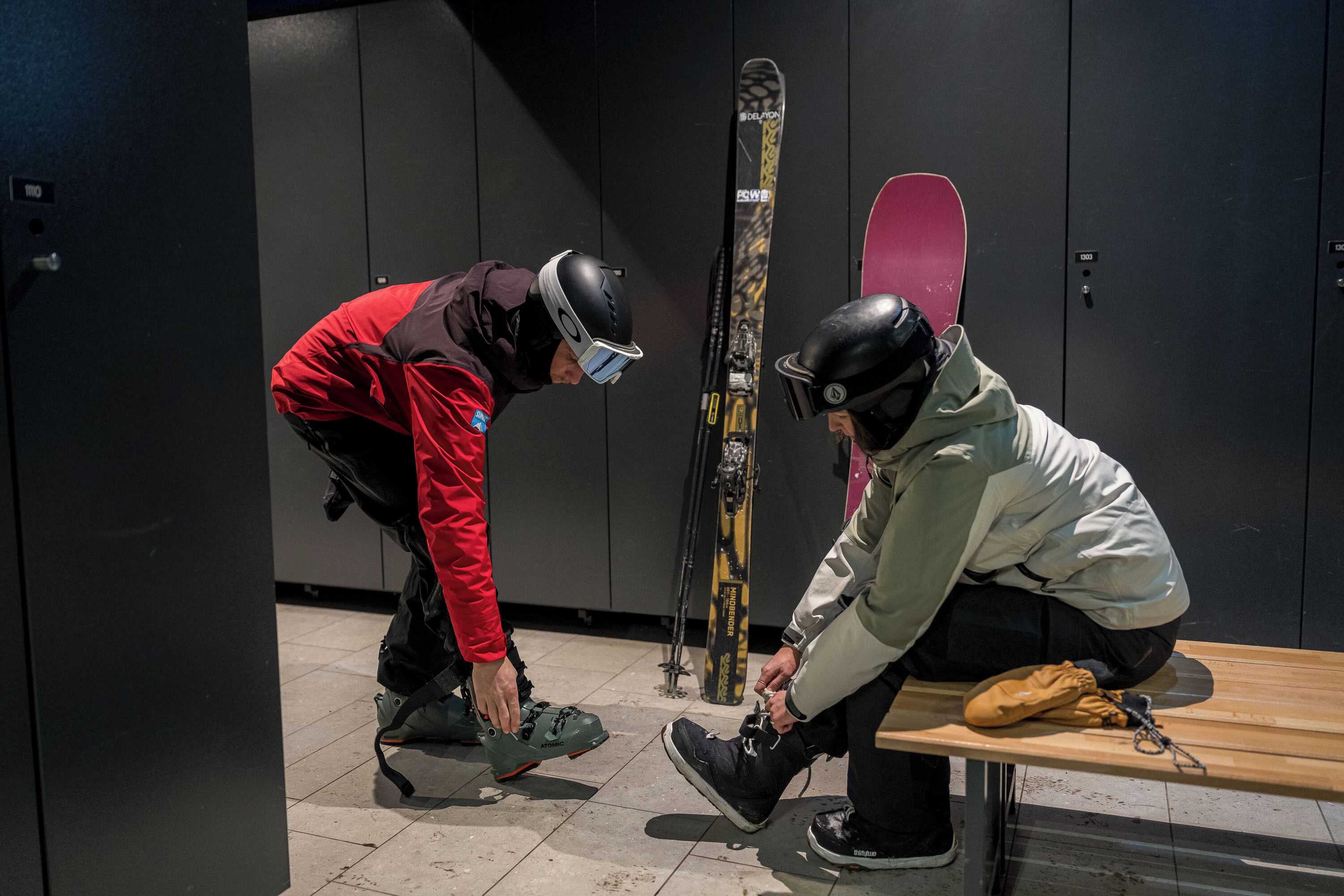 Dos personas ajustando sus botas de esquí en un vestuario, con esquíes y una tabla de snowboard pegados a la pared.