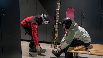 Dos personas ajustando sus botas de esquí en un vestuario, con esquíes y una tabla de snowboard pegados a la pared.