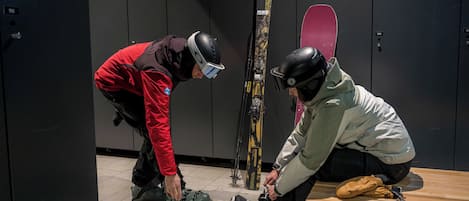 Duas pessoas ajustando botas de esqui em um vestiário com esquis e uma prancha de snowboard na parede.