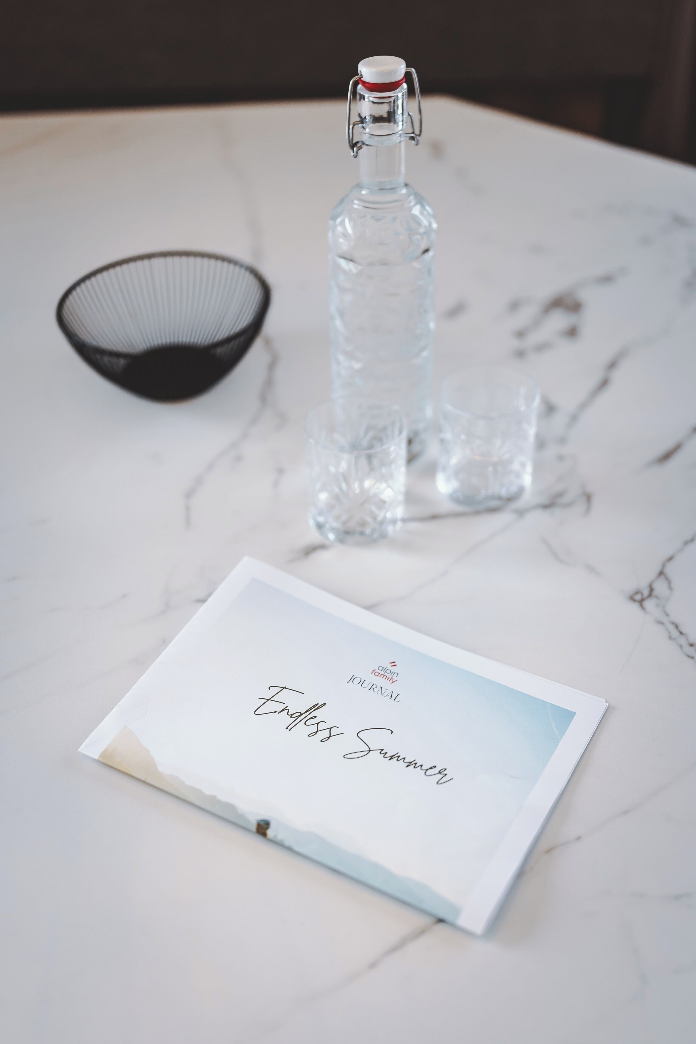 Eine klare Glasflasche mit Verschluss, ein schwarzer Drahtkorb und ein weißer Umschlag mit Schriftzug auf einer Marmorplatte.