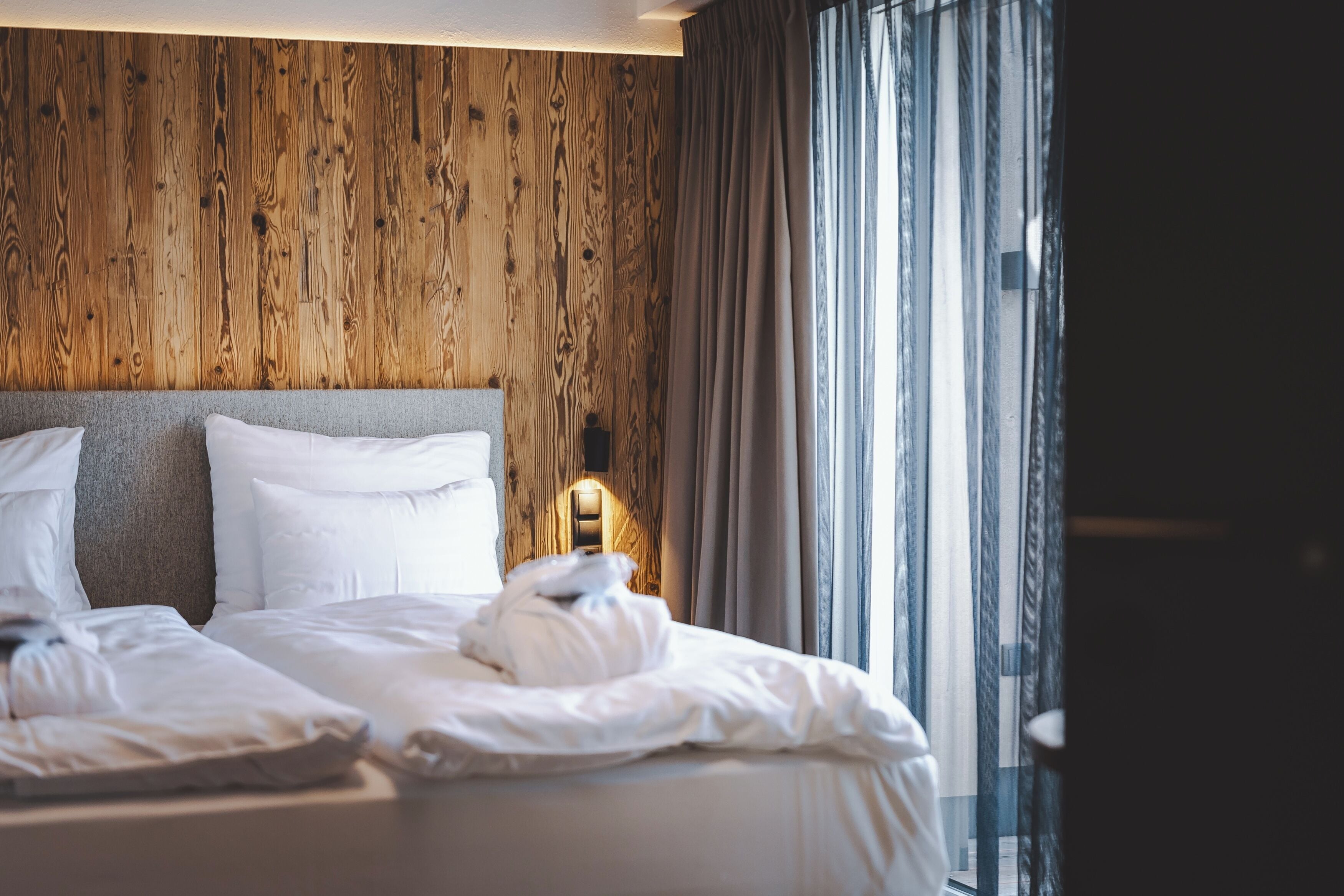Camera d'albergo con parete in legno, un ampio letto con lenzuola bianche e un comodino con una lampada.