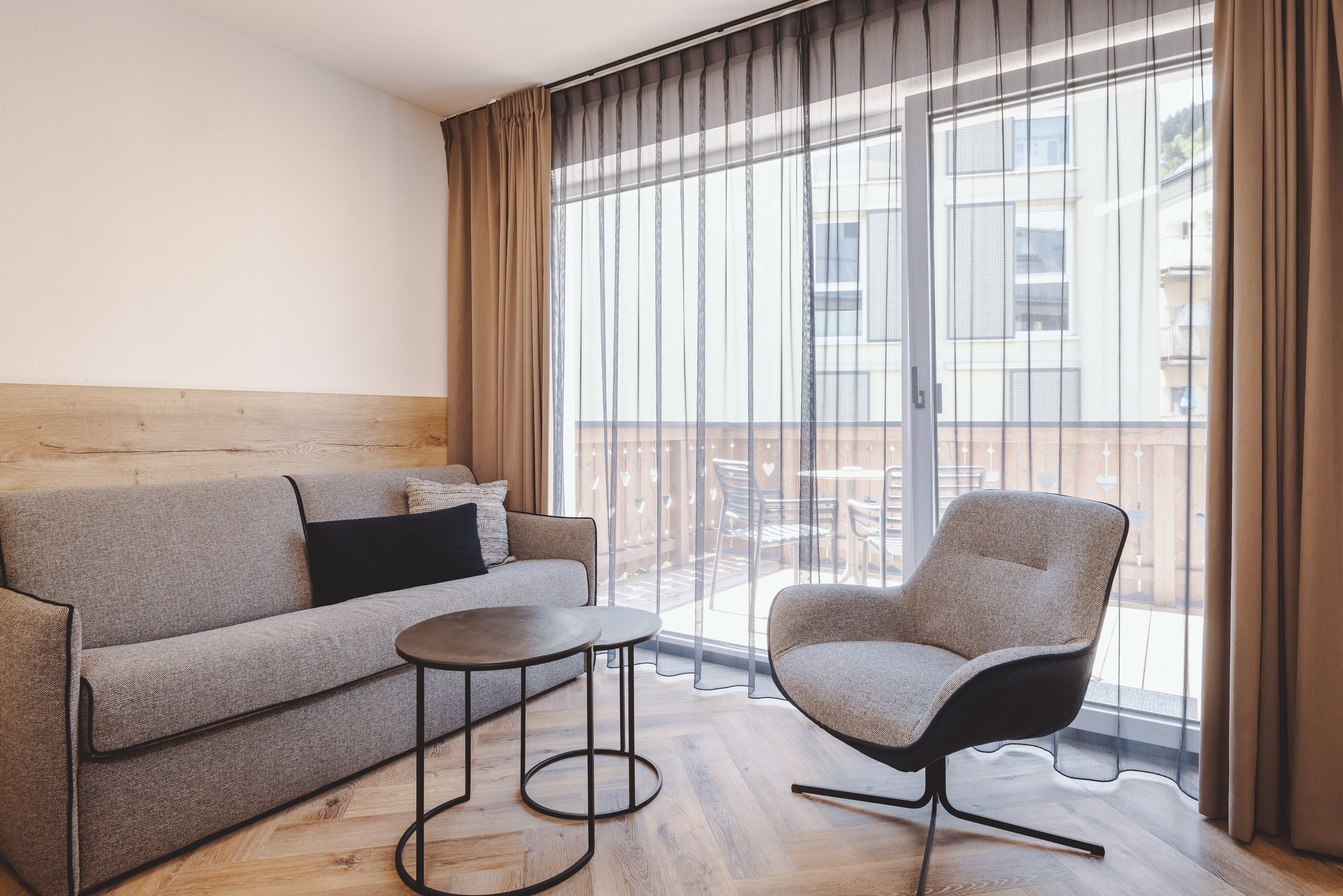 A modern living room with a sofa, a round table, and a chair, a large window with curtains, and a balcony with outdoor furniture.