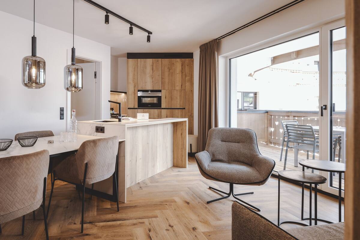 Una cocina moderna con armarios de madera, un comedor con mesa y sillas blancas, y un balcón con mobiliario de exterior.