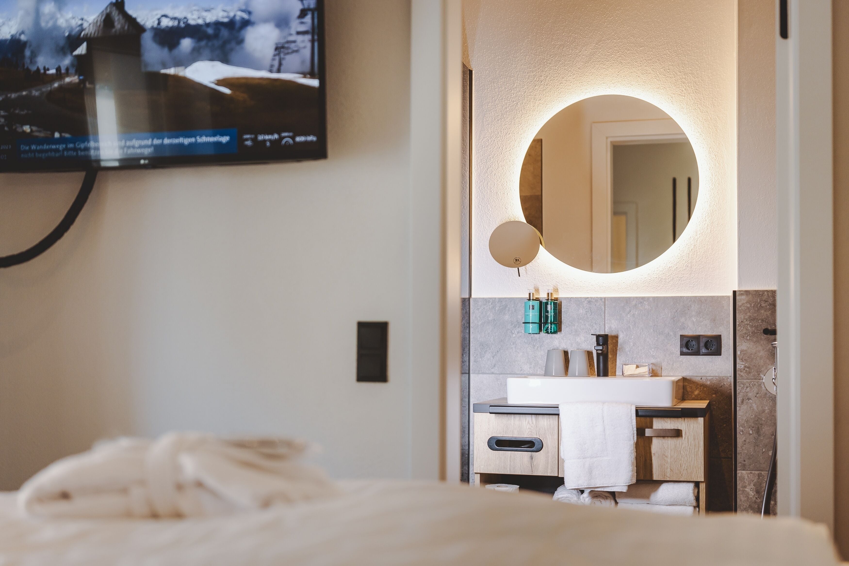 Une chambre d’hôtel moderne avec un grand lit, un miroir rond au-dessus de la coiffeuse et un téléviseur fixé au mur qui diffuse des images d’un paysage enneigé.