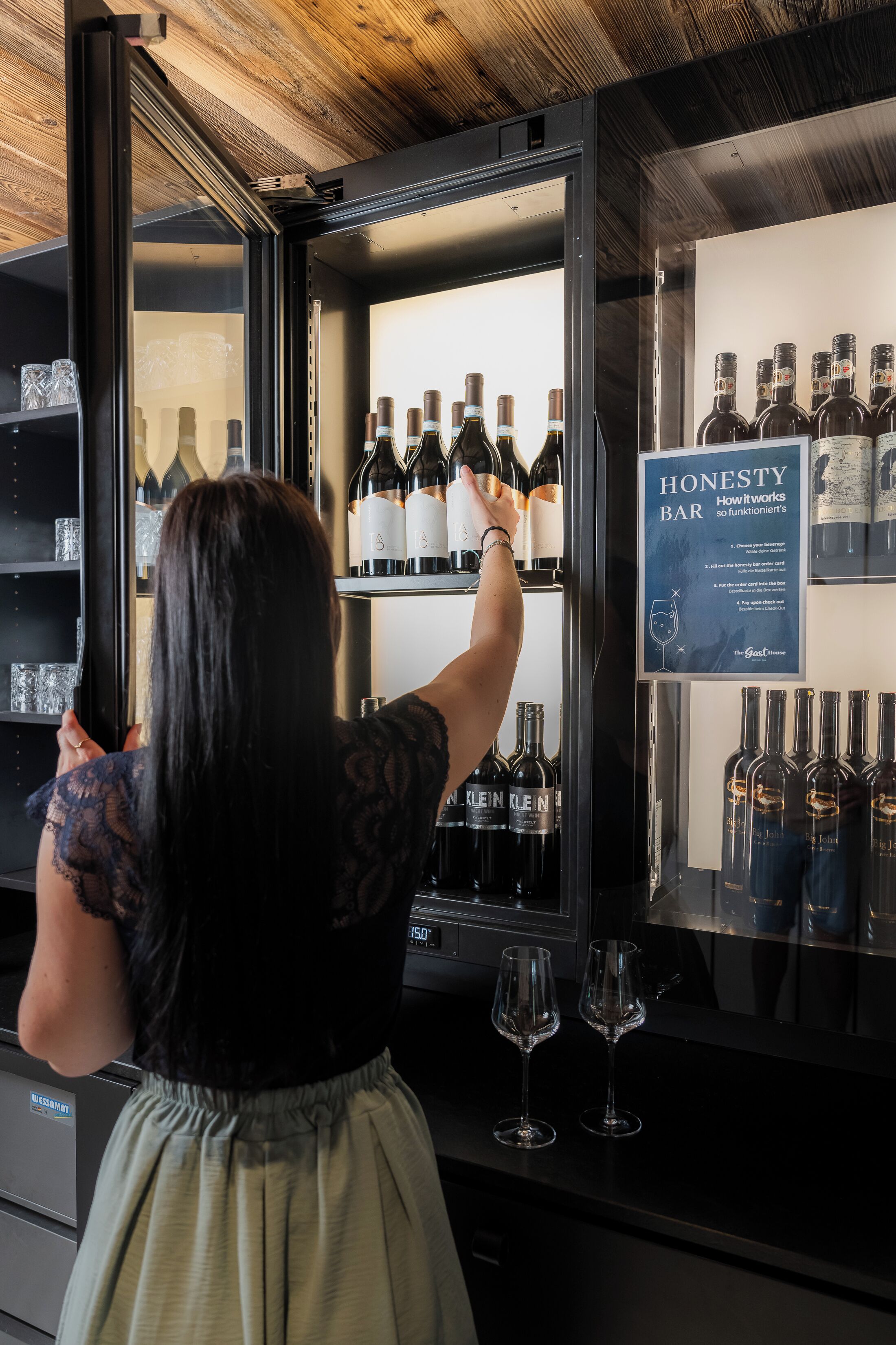 A person selecting a bottle from a wine refrigerator in a bar setting.