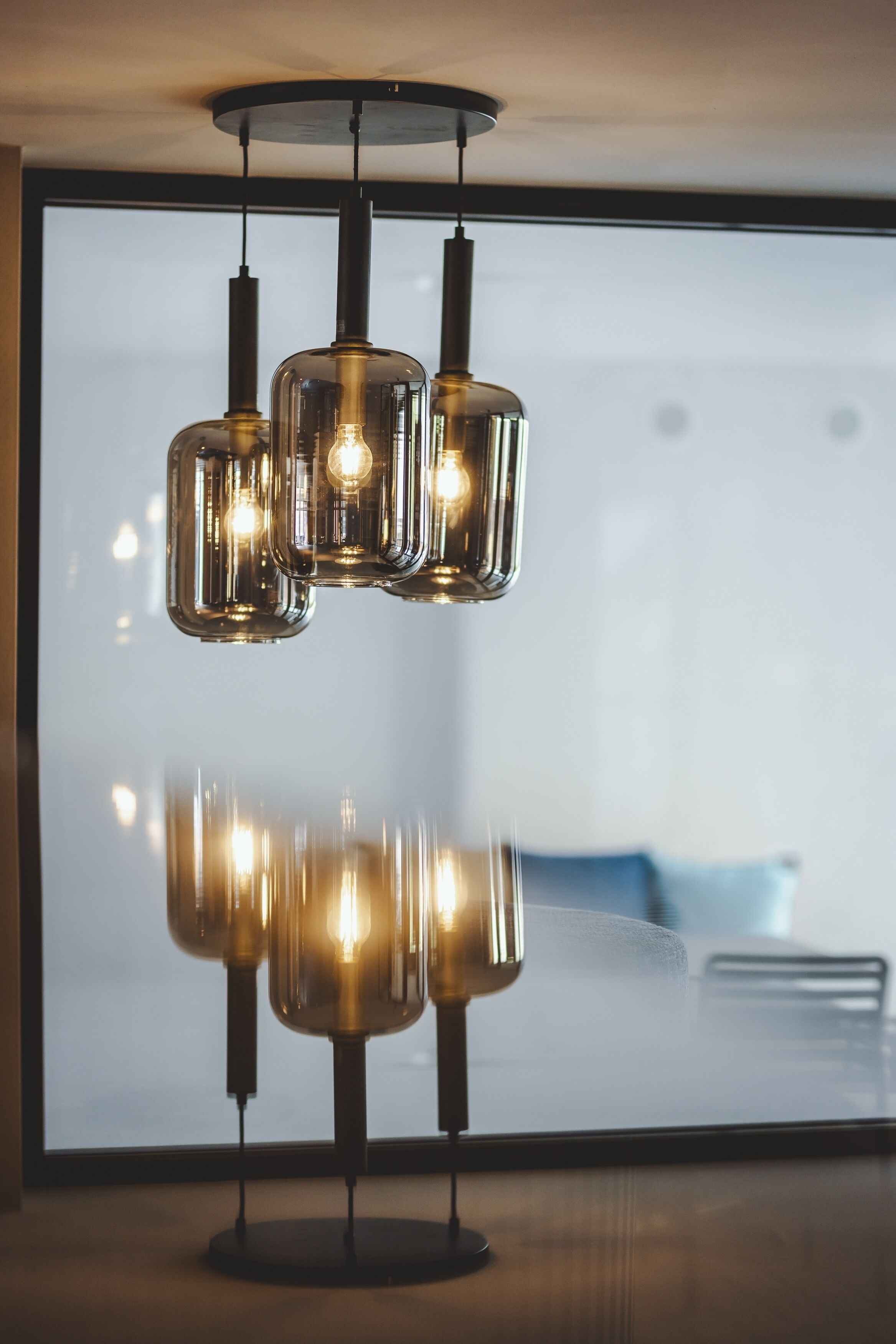 Een moderne kroonluchter met drie glazen kap en zichtbare lampen, hangend boven een glazen tafel in een kamer met een groot raam.