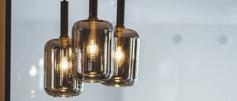 Een moderne kroonluchter met drie glazen kap en zichtbare lampen, hangend boven een glazen tafel in een kamer met een groot raam.