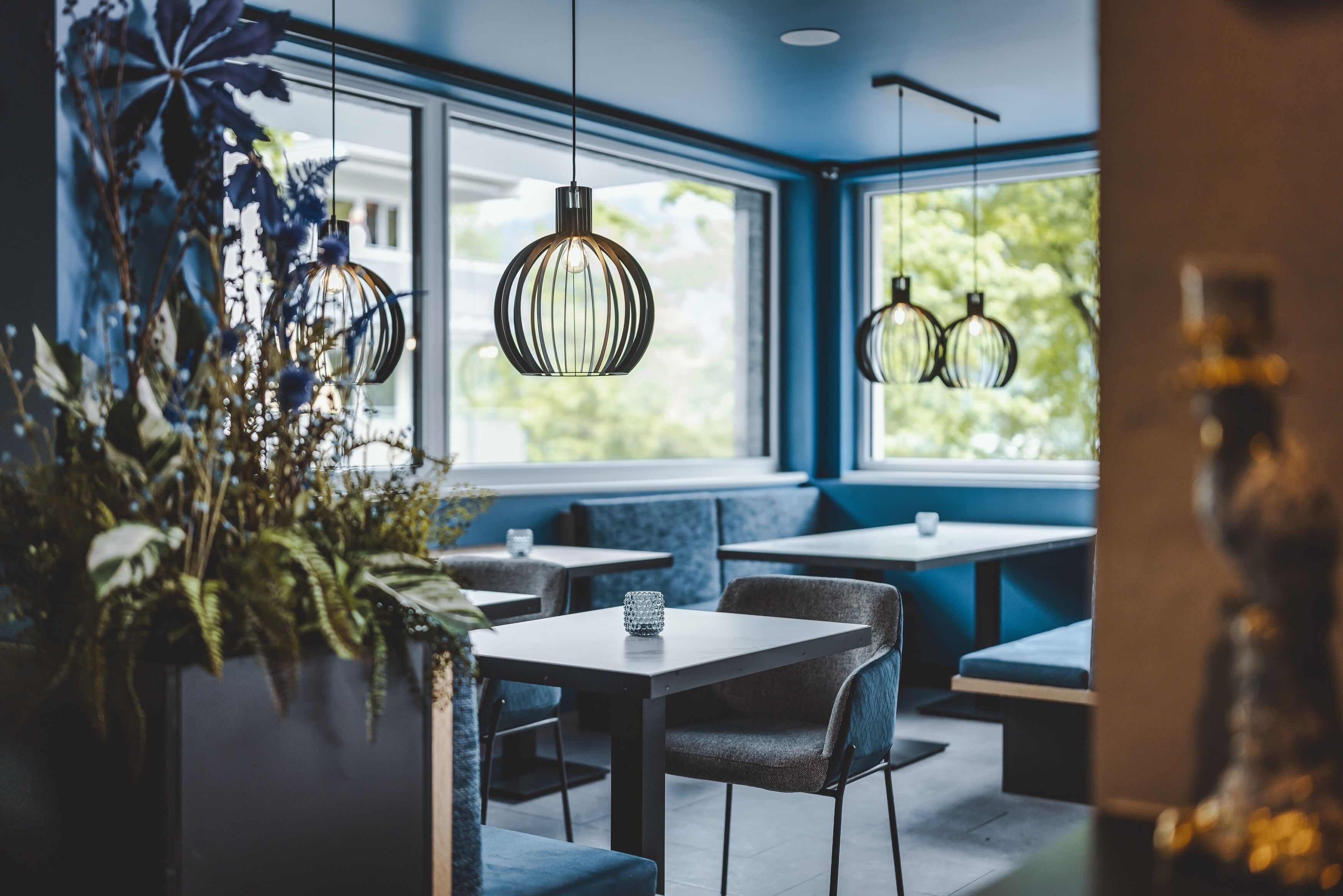 En restaurang med bord och stolar, hängande taklampor och stora fönster med utsikt över grönområden.