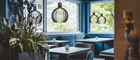 A restaurant with tables and chairs, hanging pendant lights, and large windows with a view of greenery.