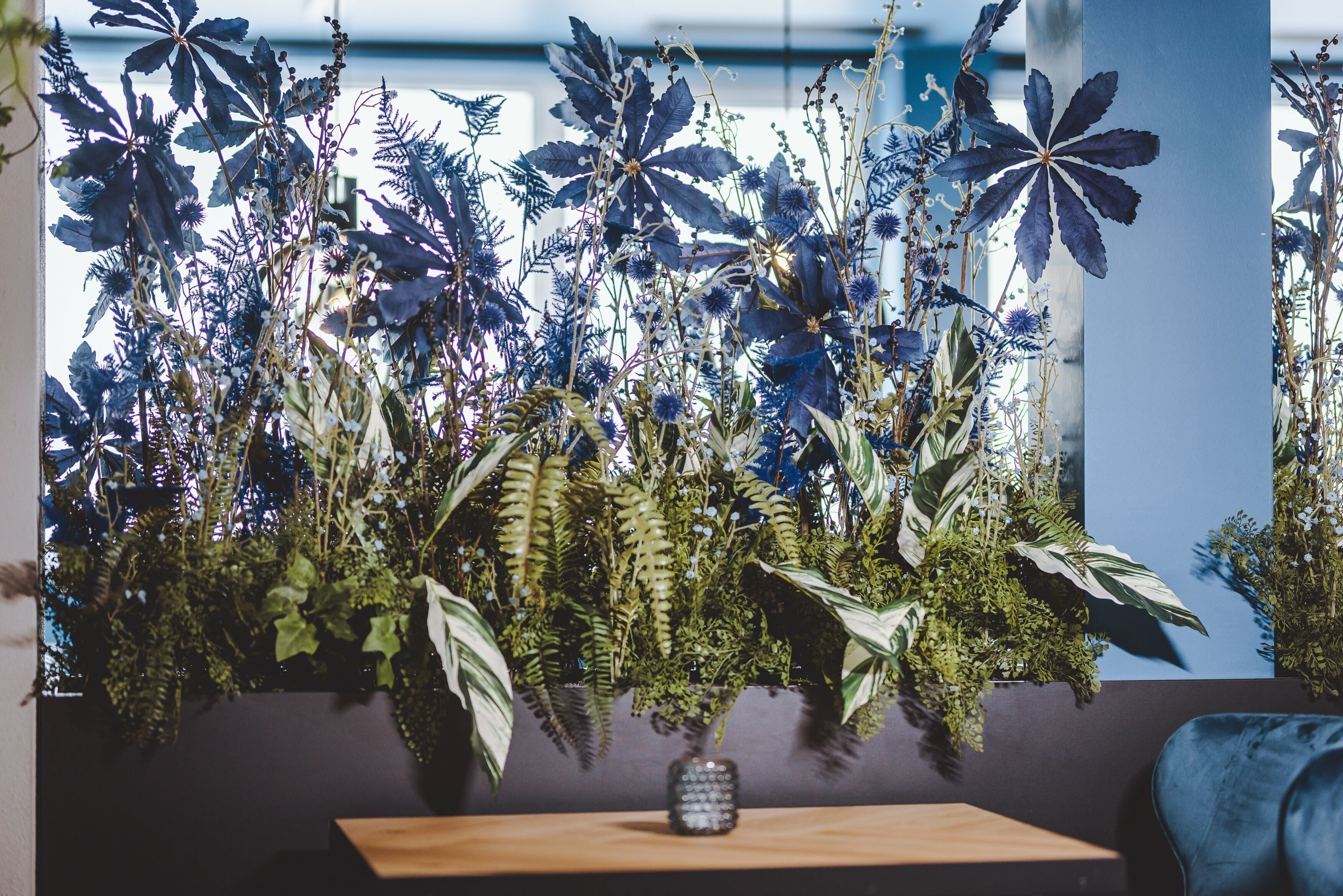 Habitación con un gran arreglo de plantas en macetas sobre una mesa de madera, un sofá oscuro y una ventana con vistas al exterior.