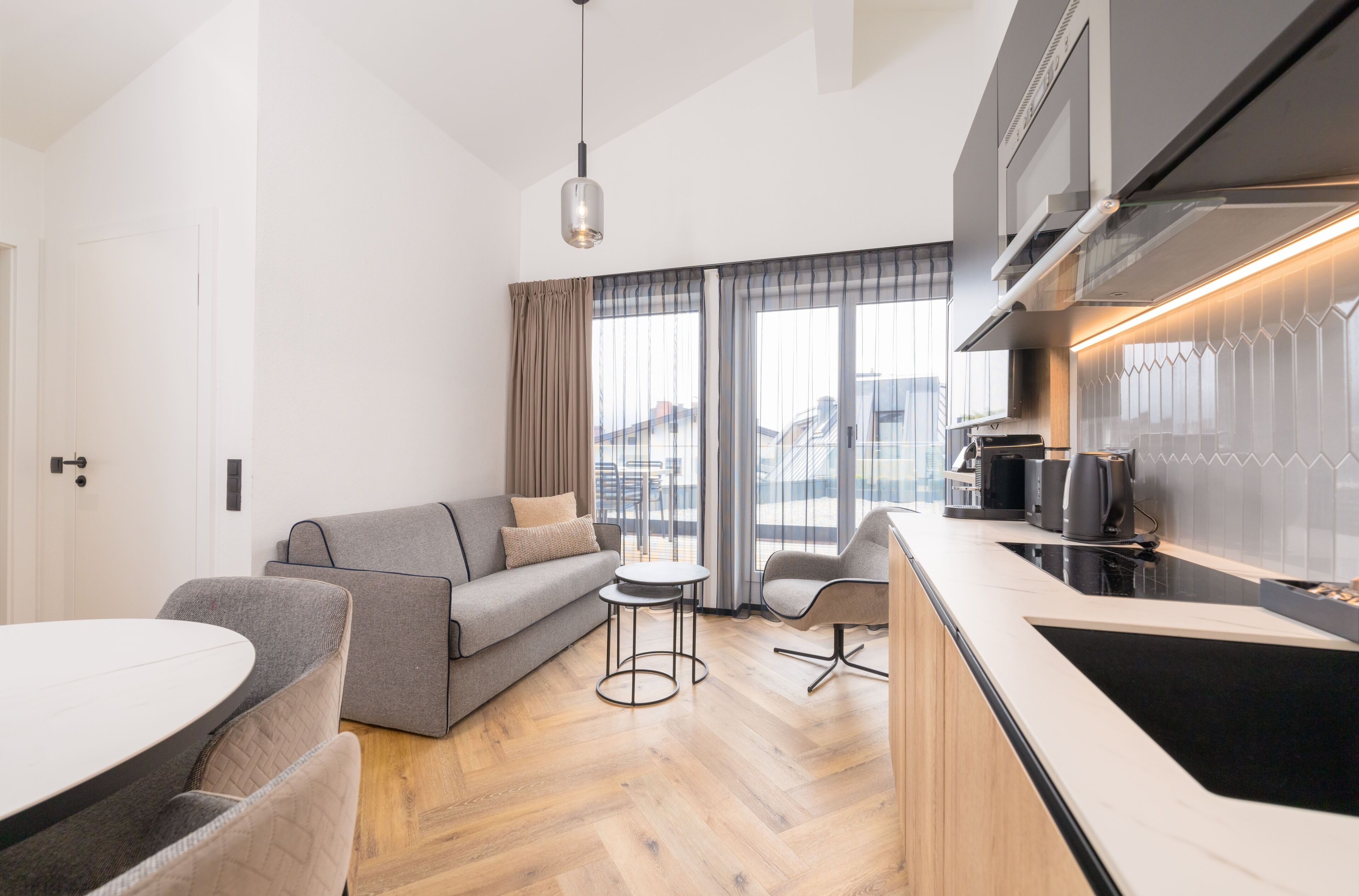 Een moderne woonkamer met een bank, fauteuils, een kleine tafel en een keukenhoek met een fornuis en kastjes.