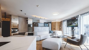 Ein modernes Wohnzimmer mit einer großen Couch, Hocker und einem Sessel. Ein Fernseher ist an der Wand befestigt, der Boden ist aus Holz und ein großes Fenster bietet einen Blick auf die Stadt.