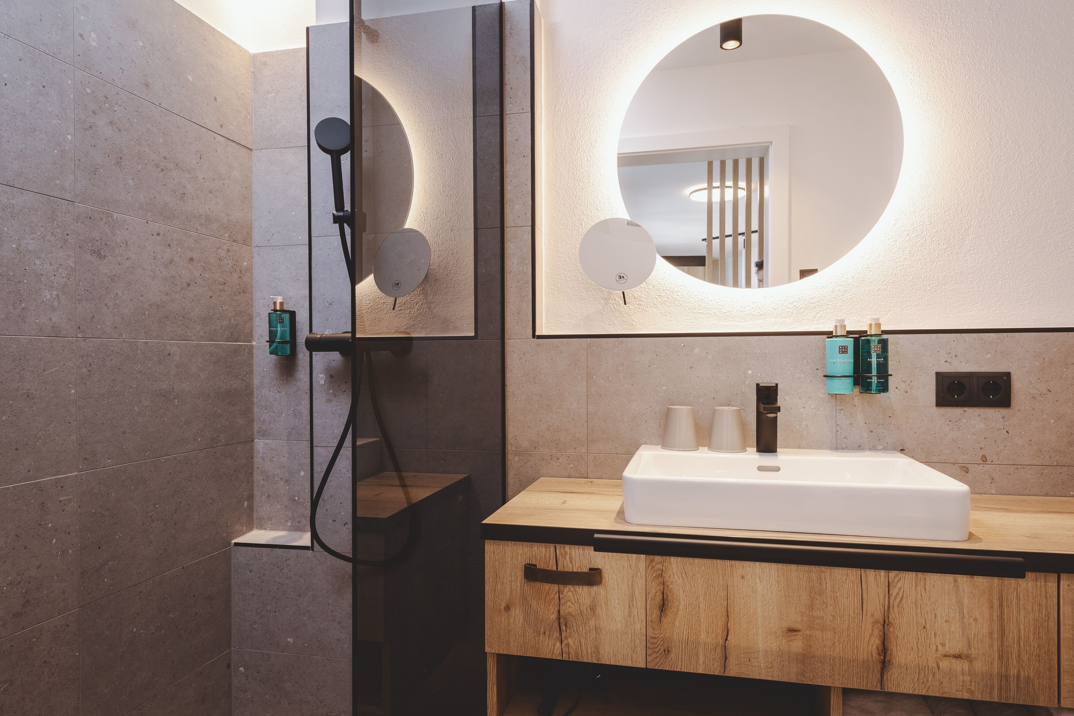 Un baño moderno con tocador de madera, un espejo grande con iluminación circular y una zona de ducha.