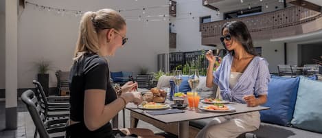 Un espace de restauration en plein air avec une table dressée pour deux personnes, entouré de chaises noires, d’un canapé orné du logo « GRANDS COUSSINS » et illuminé par des guirlandes lumineuses suspendues au-dessus.