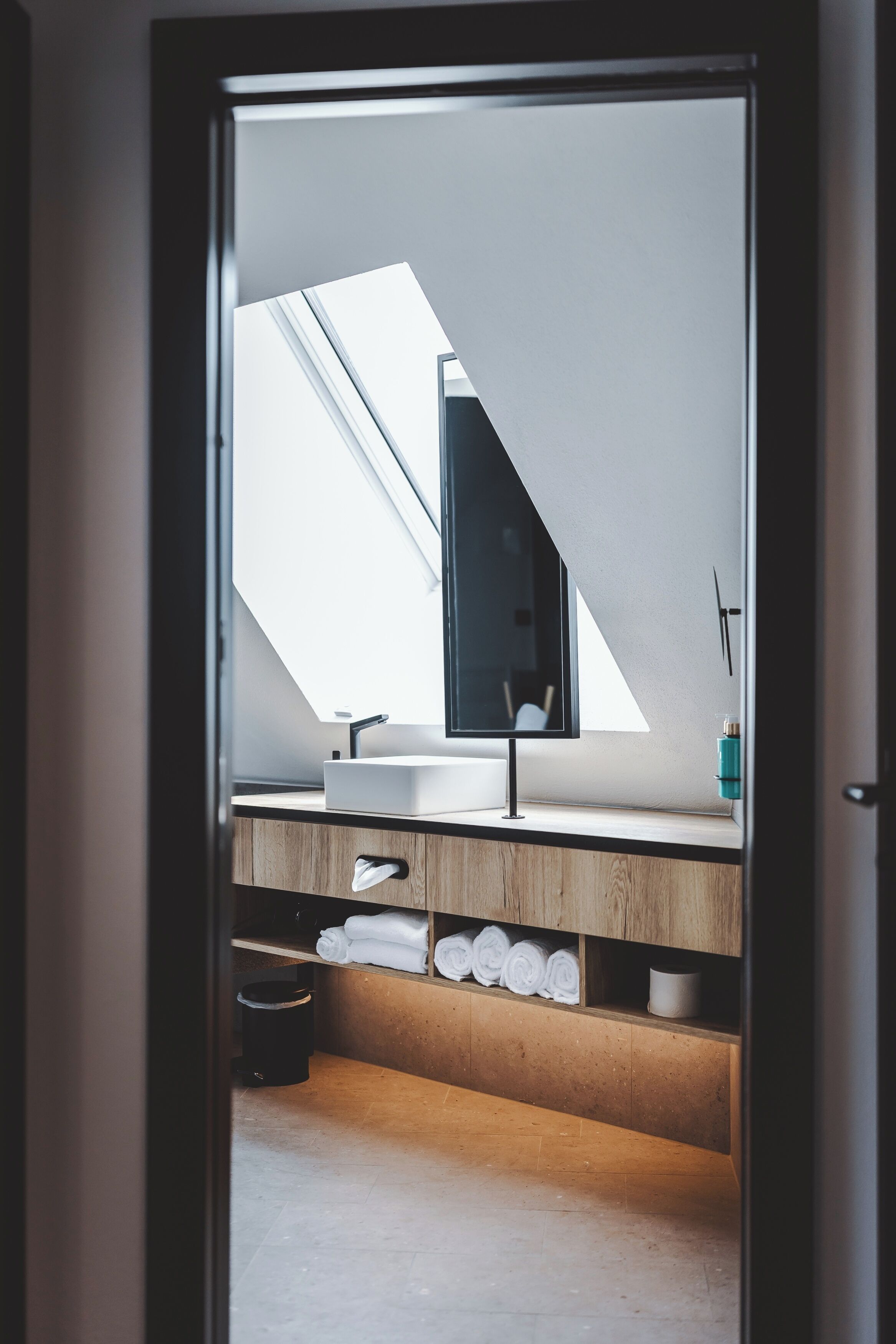 A modern bathroom with a skylight, a sink, and a mirror. There are towels and a trash can visible.