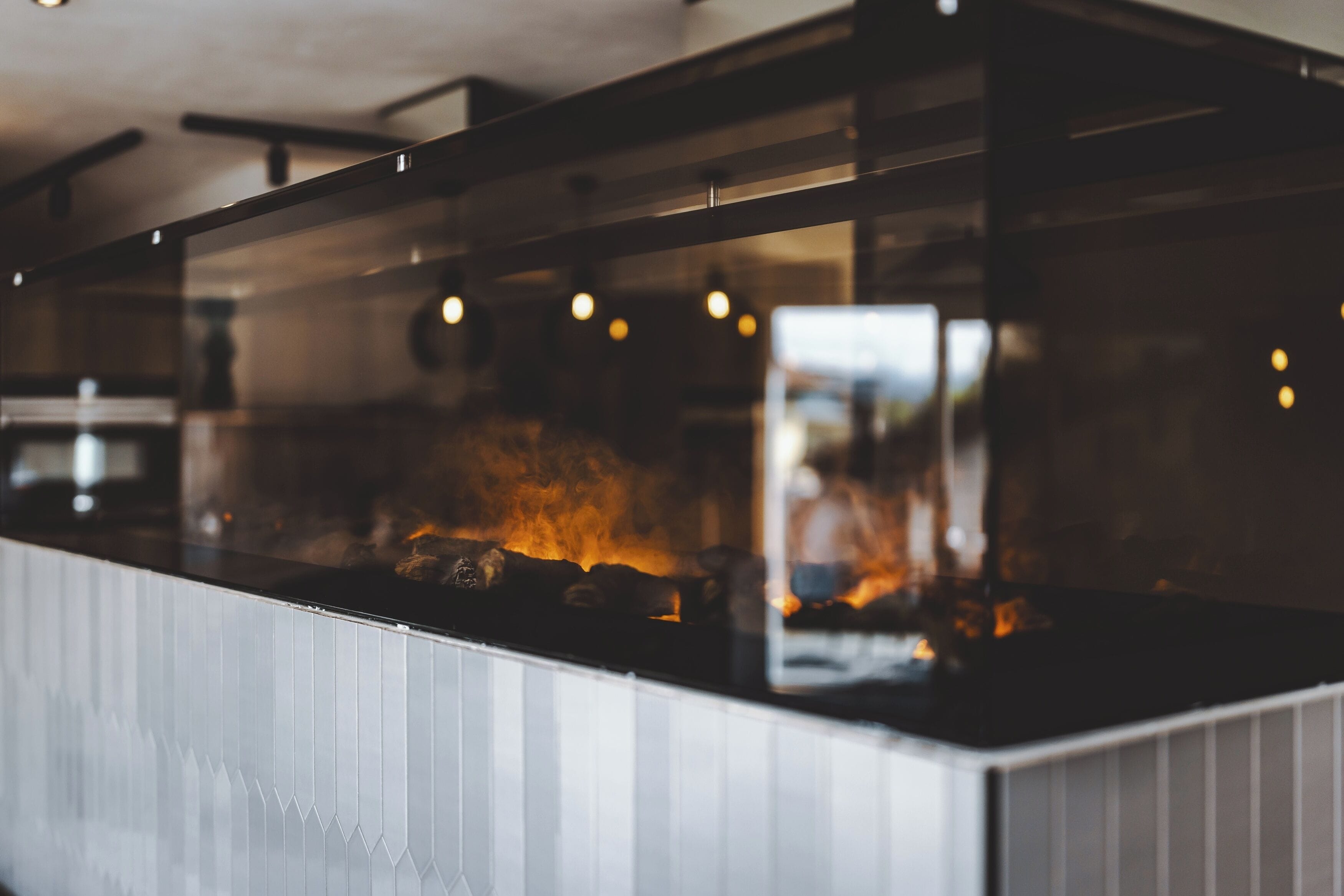 A modern kitchen with a glass partition, a visible fire, and a person in the background.