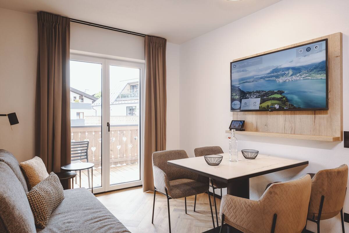 Una sala de estar moderna con sofá, una mesa de comedor con sillas, un televisor de pantalla plana en la pared y un ventanal corredero que da a un balcón.