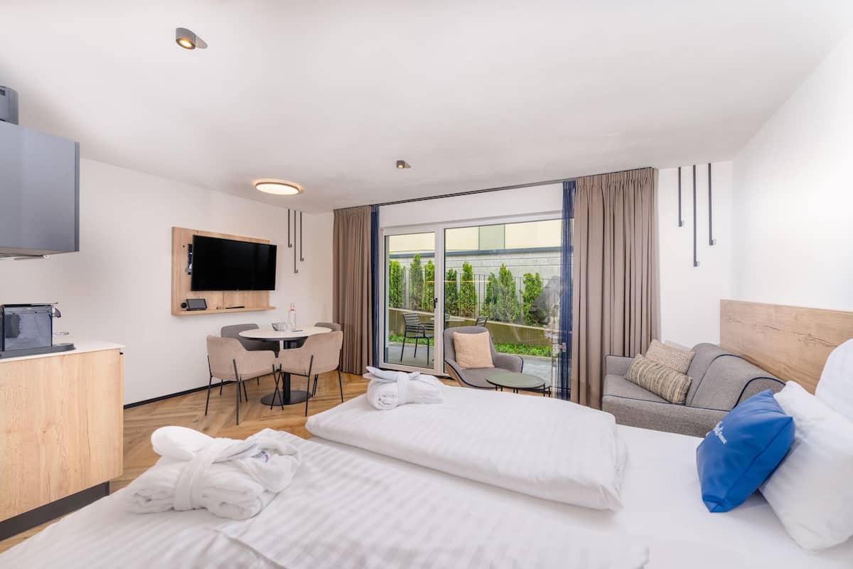 Habitación de hotel con cama, sofá, escritorio con silla, televisor y balcón con vista a zonas verdes.