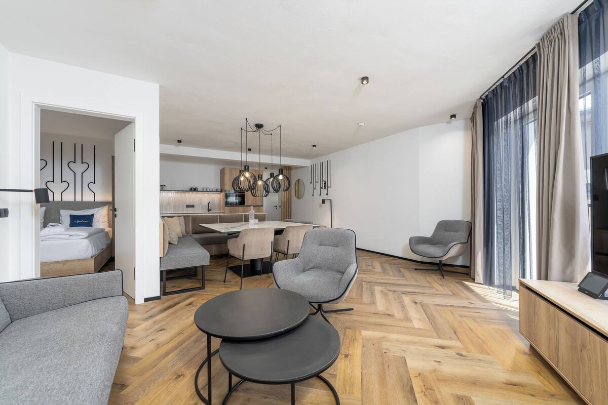 Una sala moderna con un sofá, dos mesas redondas, un comedor con una mesa y sillas, una cocina al fondo y un ventanal con cortinas.