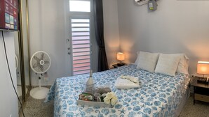A hotel room with a bed, bedside lamps, a fan, a TV, and a window with blinds.