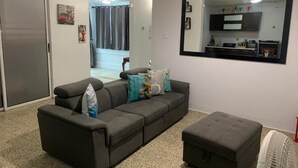 A living room with a grey sofa, a matching ottoman, a large mirror, and a white fan.