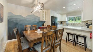 A dining area with a wooden table and chairs, a mural on the wall, and a kitchen with white cabinets and a window.