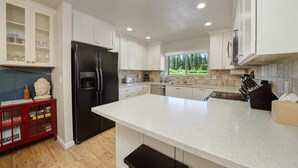 A modern kitchen with white cabinetry, a black refrigerator, a large island, and a view of a green landscape through the window.