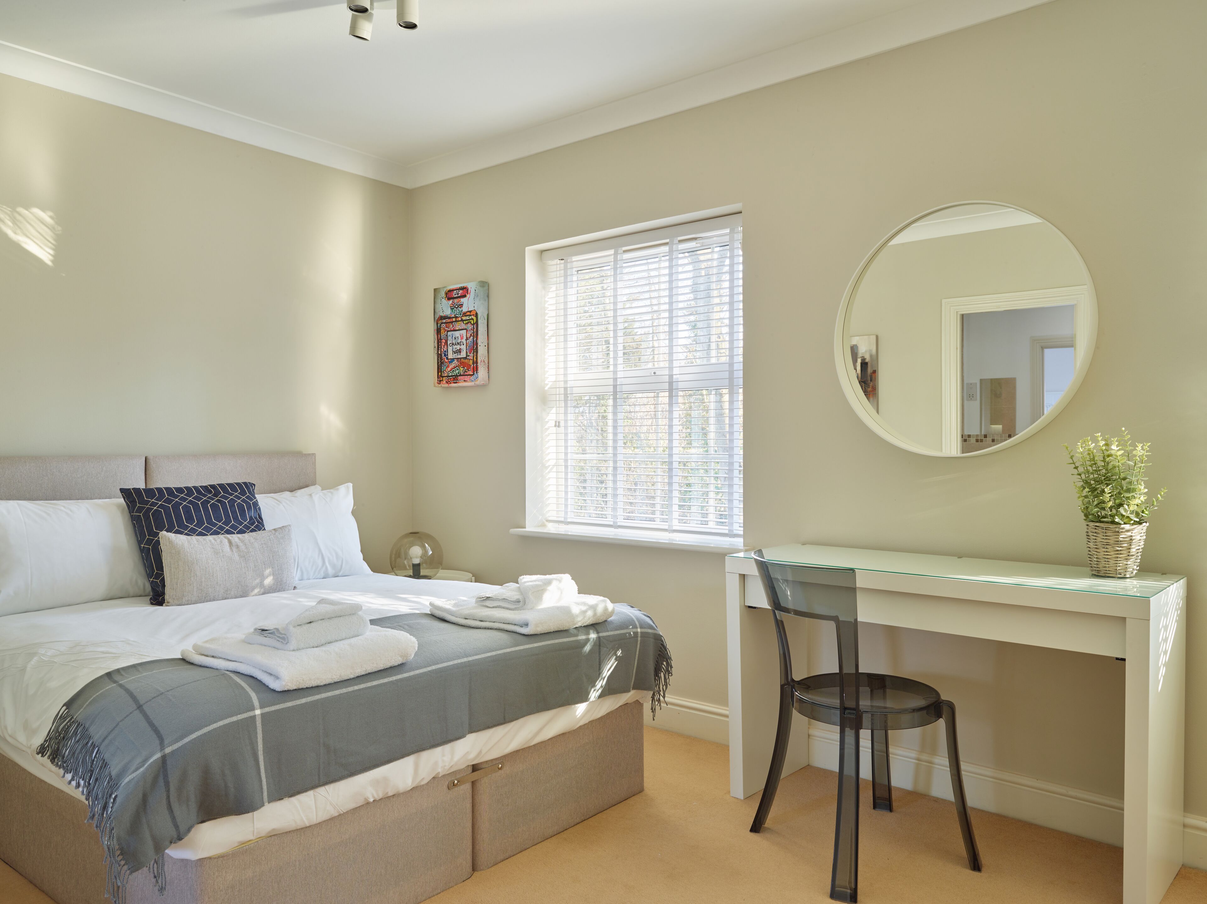 Een slaapkamer met een bed, een bureau, een stoel, een raam met zonwering, een ronde spiegel en een hangplant.