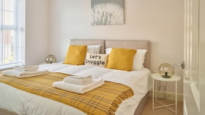 A neatly made bed with white and yellow bedding, a wall-mounted artwork, and a bedside table with a lamp. - Brookbank Close (Cheltenham)