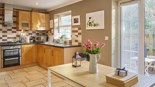 A modern kitchen with wooden cabinets, a gas stove, and a microwave. There is a window with blinds, a vase with flowers, and a table with a lantern and books. - Brookbank Close (Cheltenham)