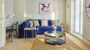 A living room with a blue sofa, a glass coffee table, and a rug. There are two side tables with lamps and a framed floral painting on the wall. - Brookbank Close (Cheltenham)
