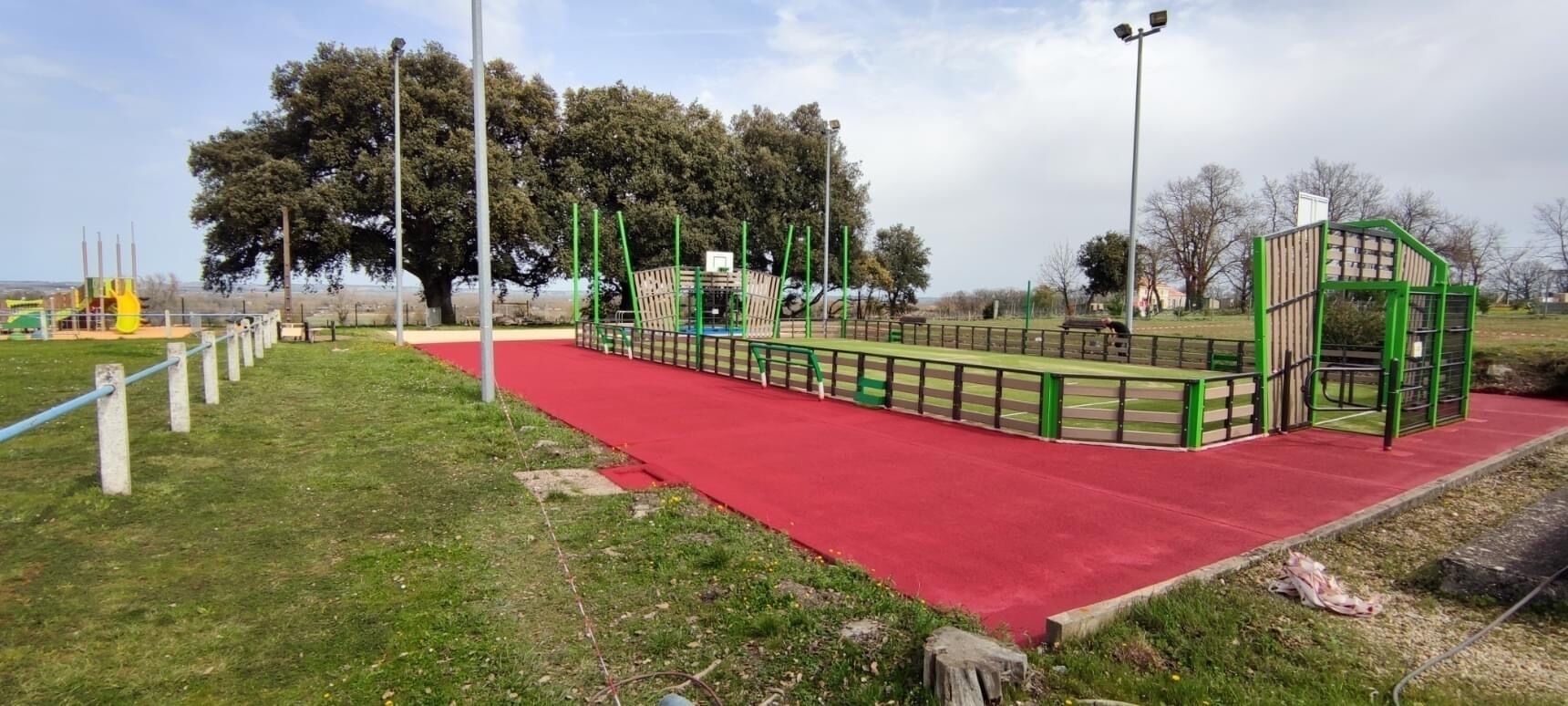 A playground with a red rubber surface, green play structures, and a large tree in the background.