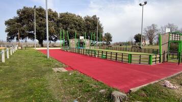 A playground with a red rubber surface, green play structures, and a large tree in the background.