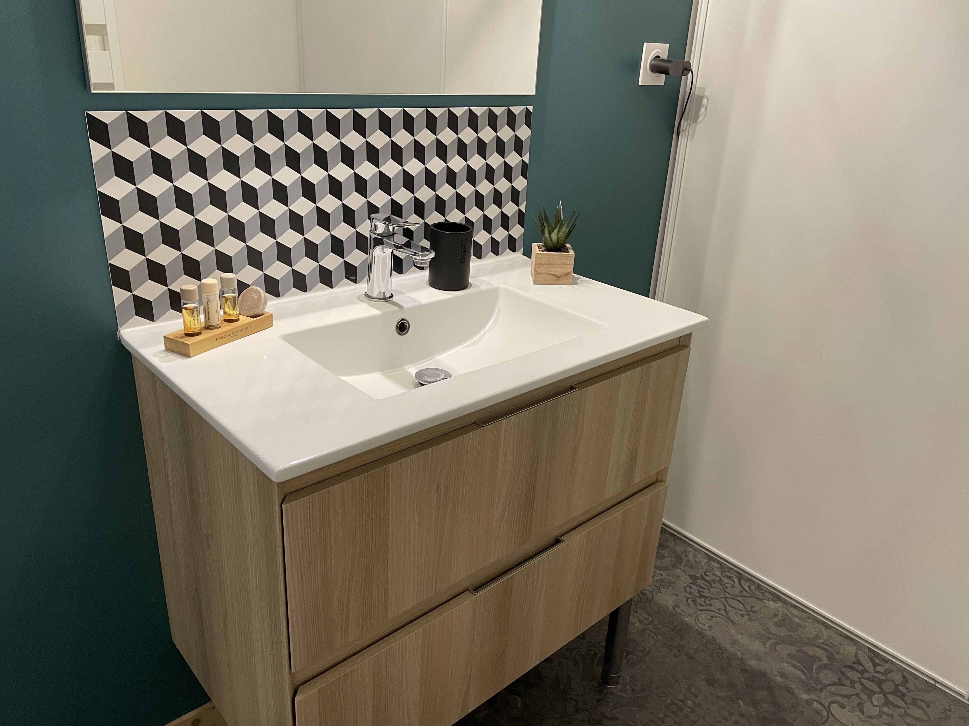 Ein Badezimmer mit einem weißen Waschbecken, einem Holzkosmetikschrank und einer geometrisch gemusterten Rückwand.