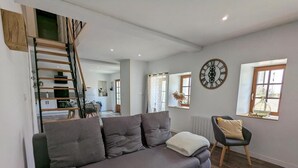 A modern living room with a grey sofa, a wooden staircase, a dining area, and a large wall clock.