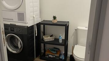 A compact bathroom with a washing machine, a small shelf with toiletries, and a toilet.