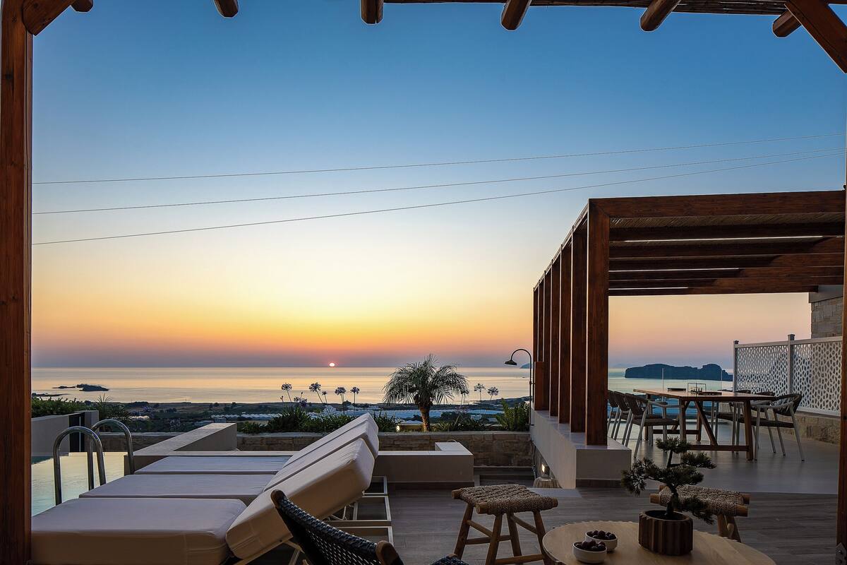 Eine Terrasse mit Liegestühlen und Esstisch mit Blick auf das Meer bei Sonnenuntergang.