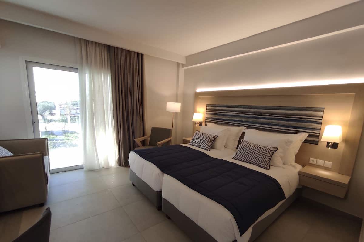 A modern hotel room with a large bed, two bedside tables with lamps, a chair, and a view of the outdoors through a large window.
