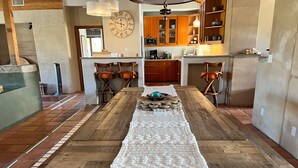 A spacious dining area with a long wooden table, chairs, and a white runner. A kitchen is visible in the background with wooden cabinets and a clock. - Majestic House with Unique Architecture, Forever Views, Pool, Spa, and Game Room (Borrego Springs)