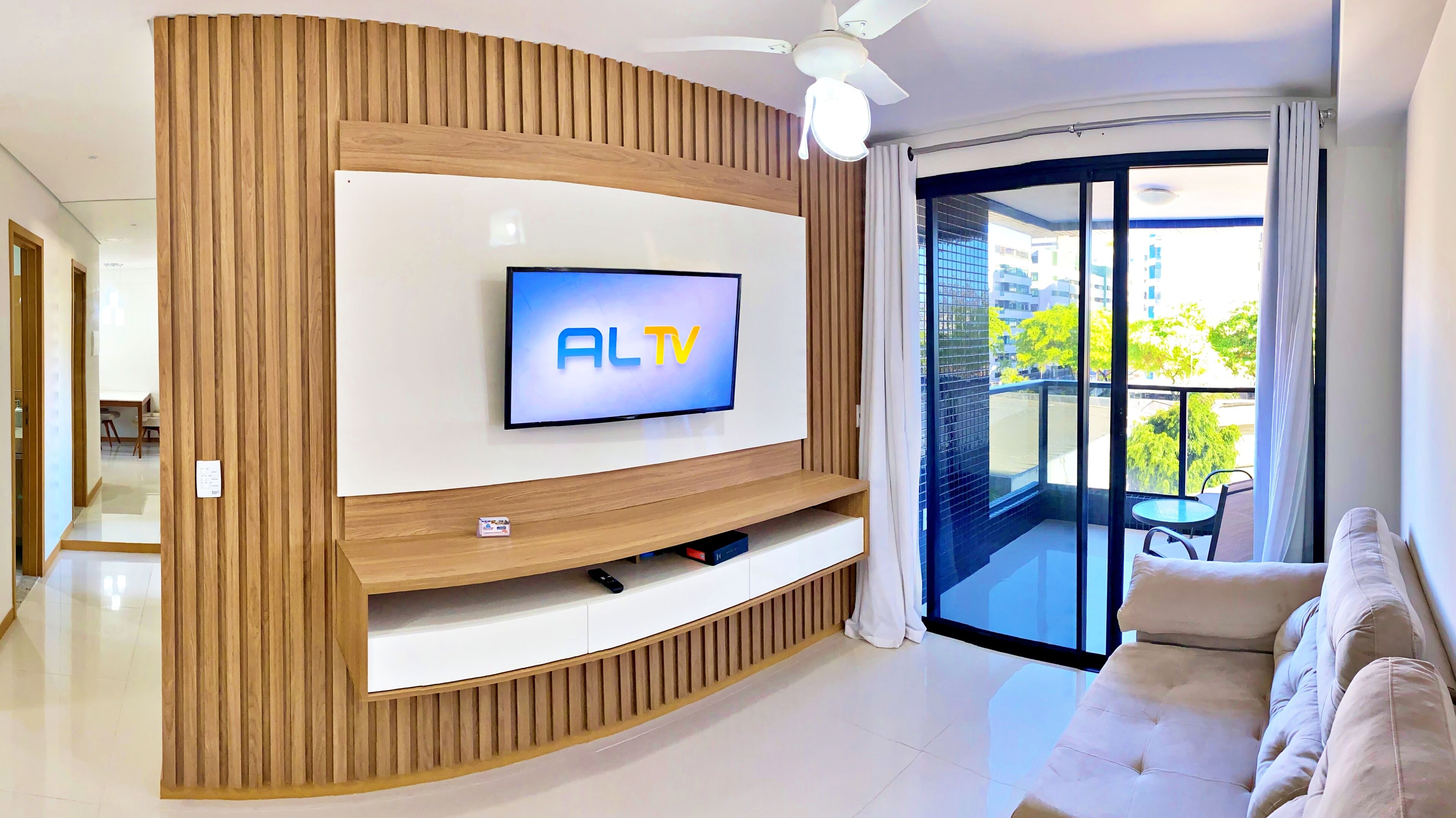 A modern living room with a flat-screen TV mounted on a wooden accent wall, a sofa, and a balcony with a glass door.