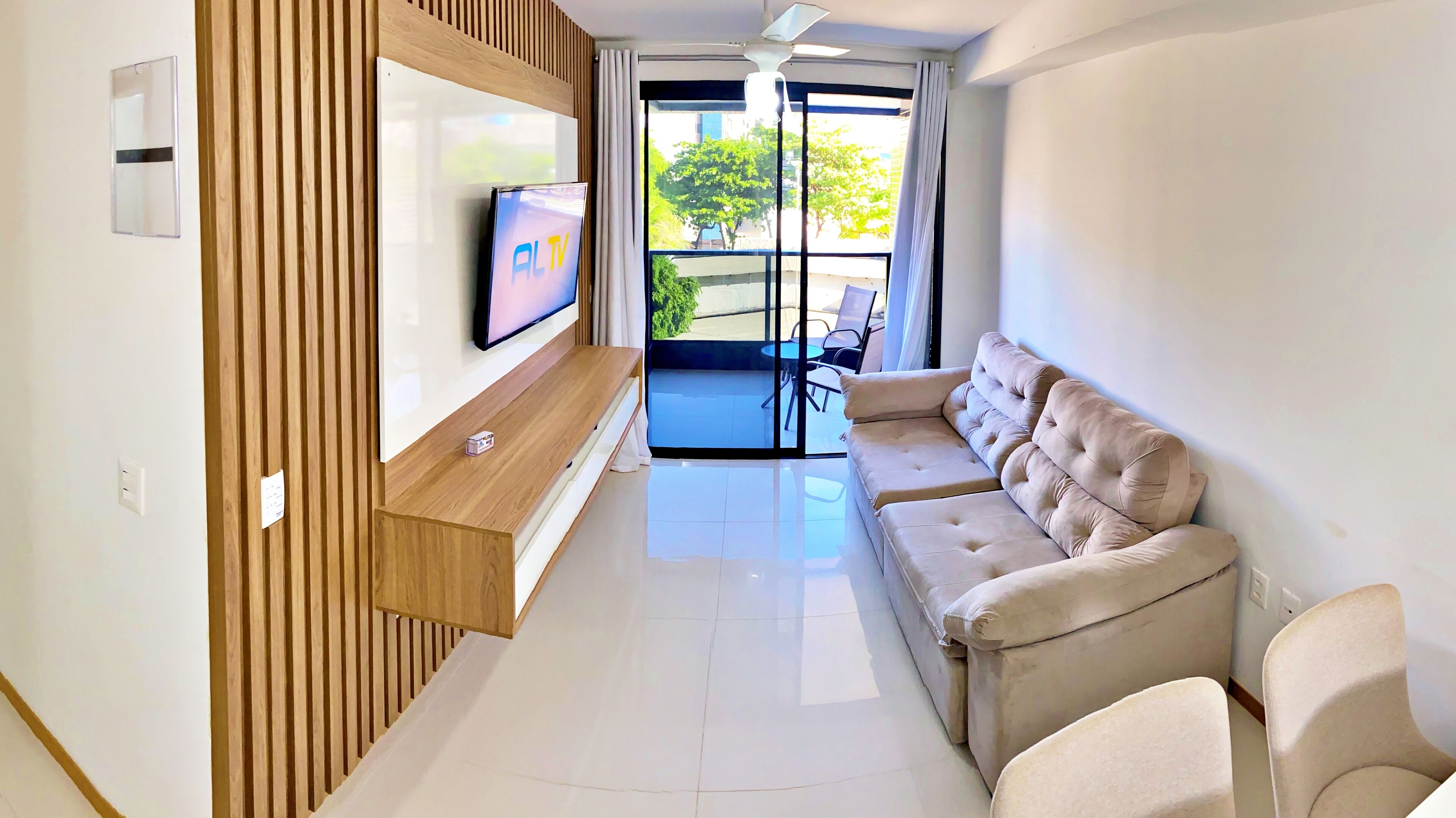 Una sala de estar moderna con un televisor de pantalla plana montado en un panel de madera, un sofá color beige y un ventanal que da a un área exterior.