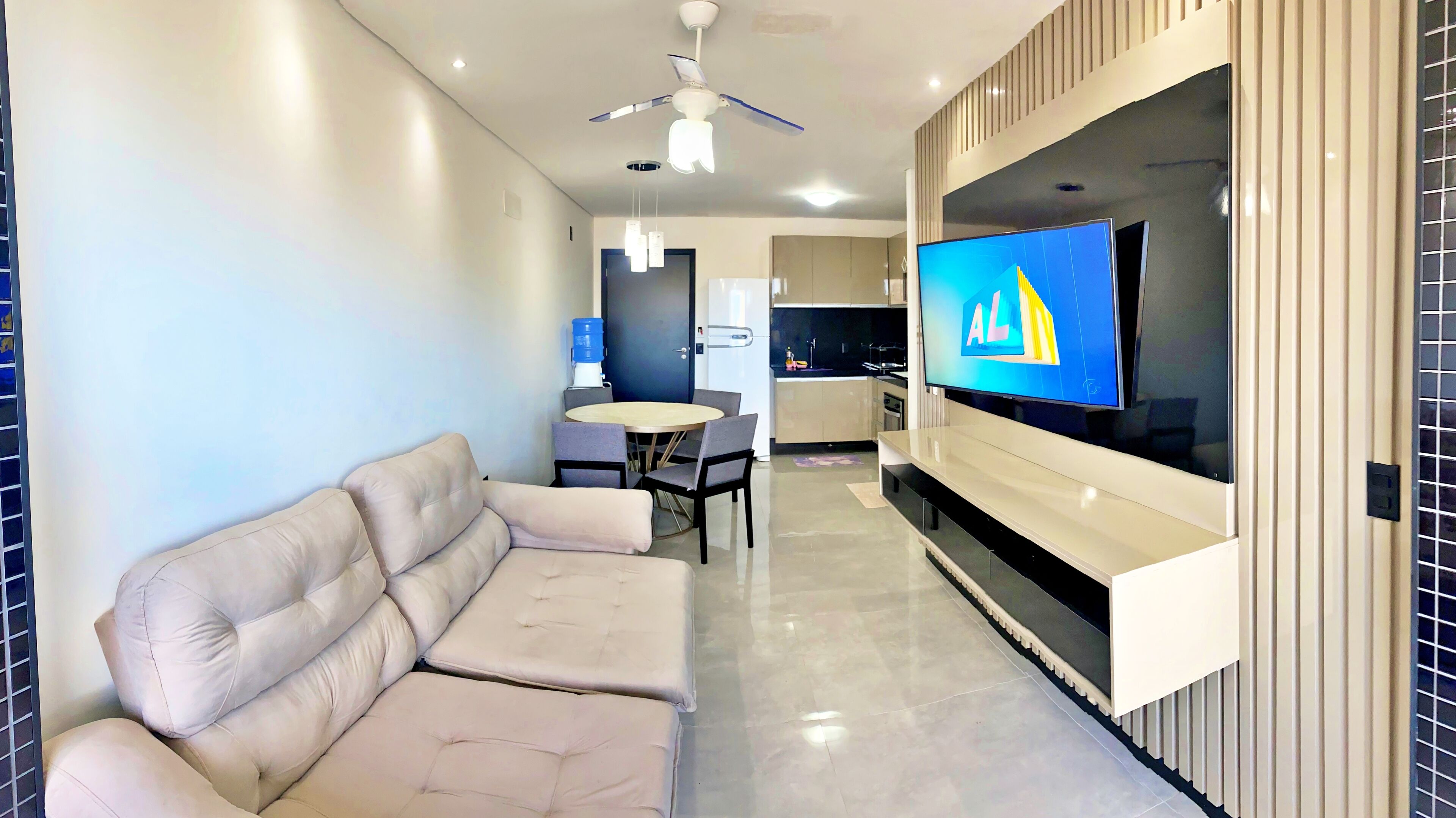 A modern living room with a beige sofa, a dining table with chairs, a kitchen area, and a TV mounted on the wall.