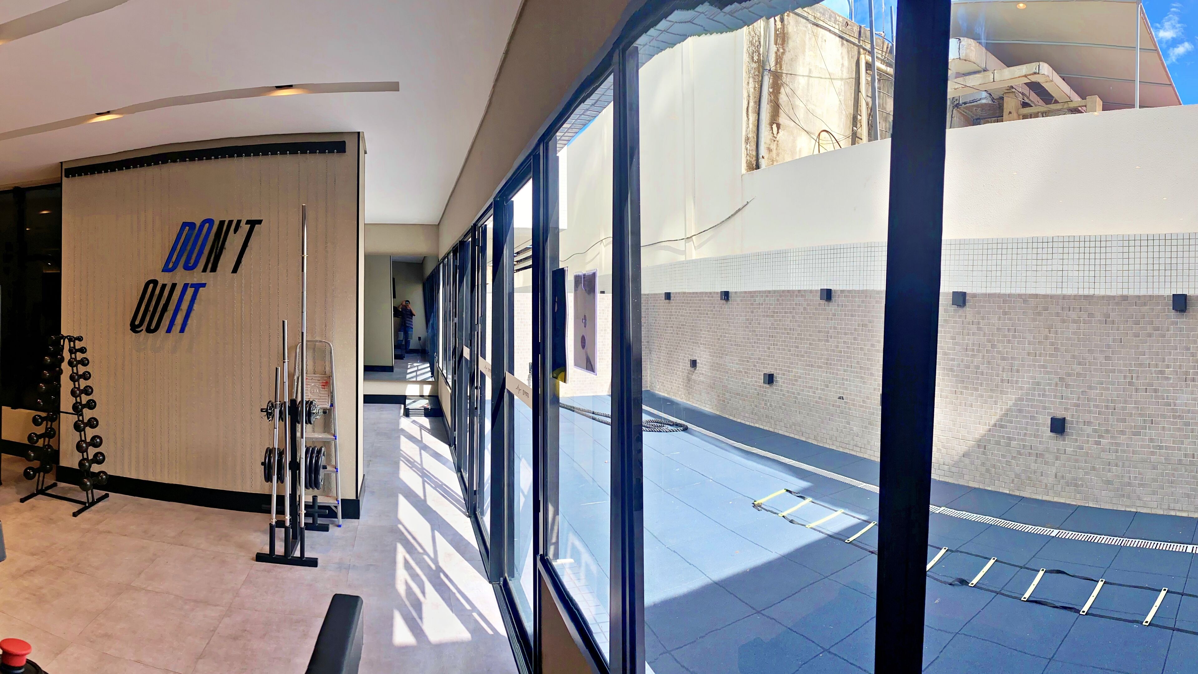 Un gimnasio con equipo de levantamiento de pesas, un letrero motivacional y grandes ventanales que ofrecen vistas del interior del edificio.