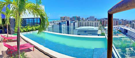A rooftop infinity pool with a city view, red lounge chairs, and a palm tree.