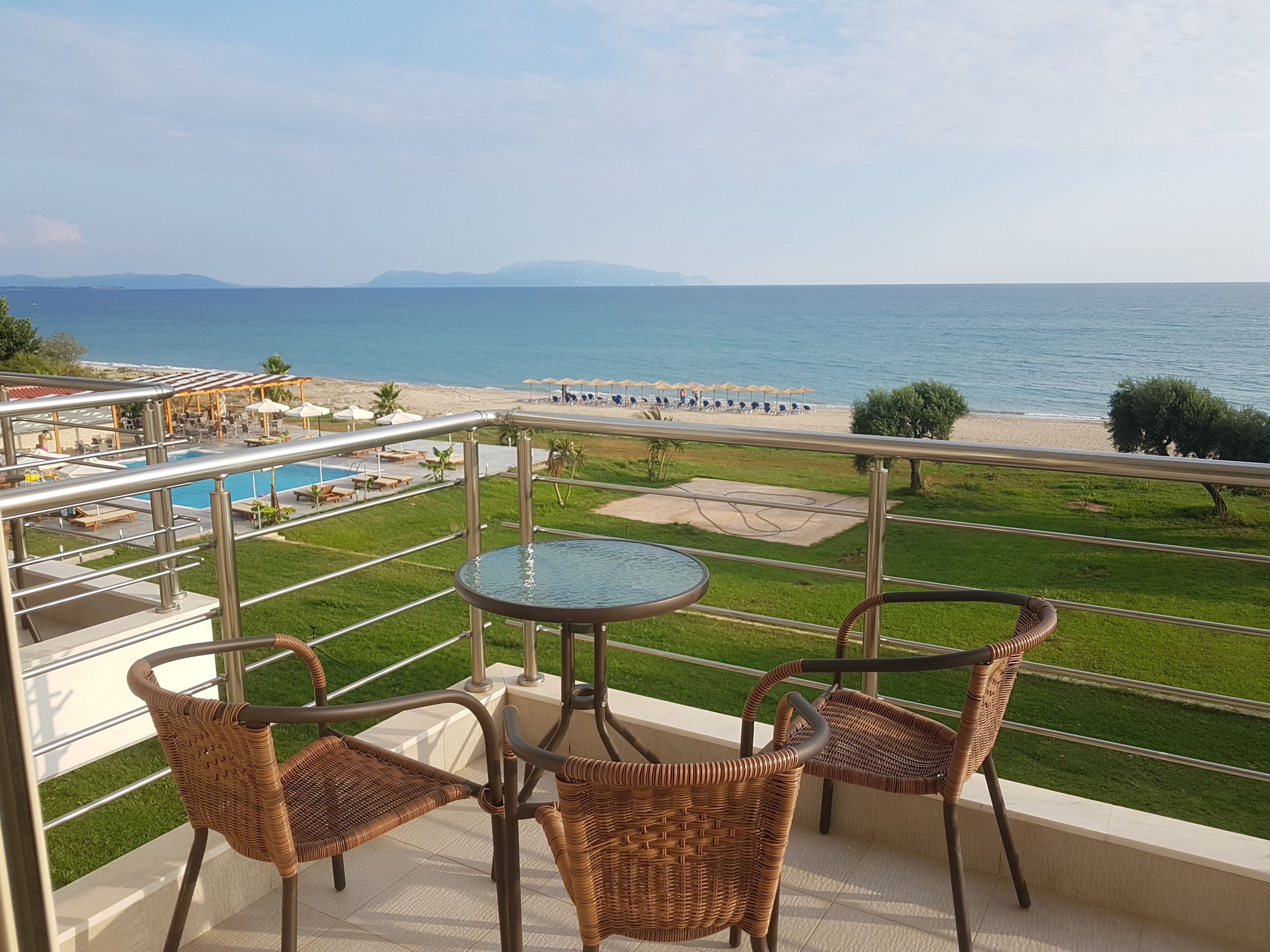 A balcony with wicker chairs and a table overlooking a pool and beach.