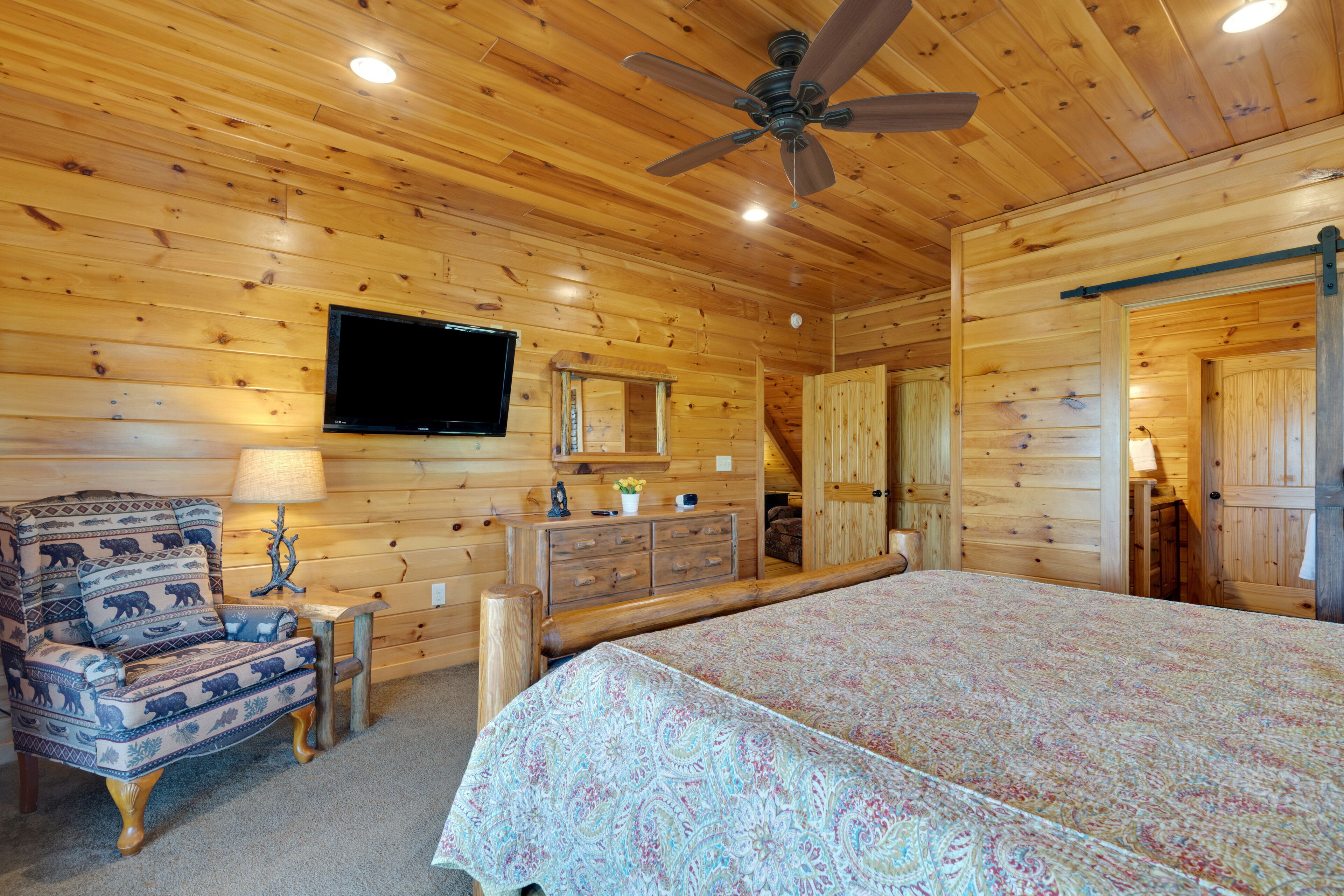 A wooden cabin bedroom with a bed, a chair, a dresser, a television, and a ceiling fan.