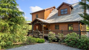 A log cabin with a porch, a stone chimney, and a gravel driveway.