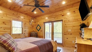 A log cabin bedroom with a bed, ceiling fan, wooden walls, and a large window.