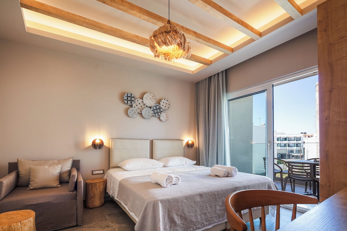 Habitación de hotel moderna con una cama grande, un sofá, una silla de madera y un balcón con vistas a la ciudad.